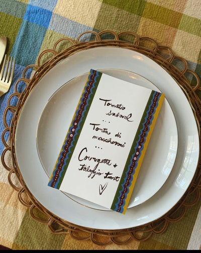 Picture of a blue, yellow and burgundy bordered menu card sitting on top of a dinner plate with handwritten menu